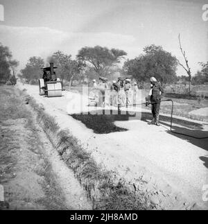 1950s, historisch, ein Arbeiter, der Wasser auf neuen Asphalt sprüht, der auf einem Teil einer unbefestigten Landstraße liegt, mit einer Dampfwalze, die darauf wartet, hinüber zu gehen und sie zu glätten, Indien. Stockfoto