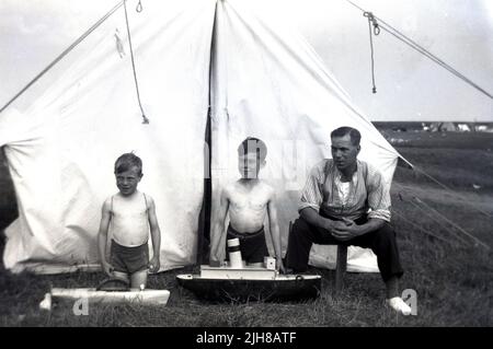 Um 1940s, historischer Camping-Urlaub auf einem Feld, knieten zwei kleine Jungen vor ihrem Zelt auf dem Gras, mit ihrem Vater neben ihnen und zeigten ihre Spielzeugboote, England, Großbritannien. Stockfoto