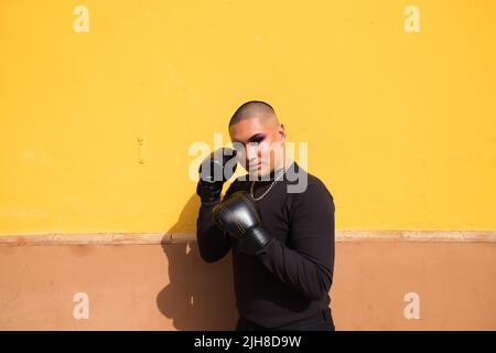 Eine junge attraktive lateinische nicht-binäre Person, die Make-up trägt und in schwarzen Boxhandschuhen an einer gelben Wand posiert Stockfoto