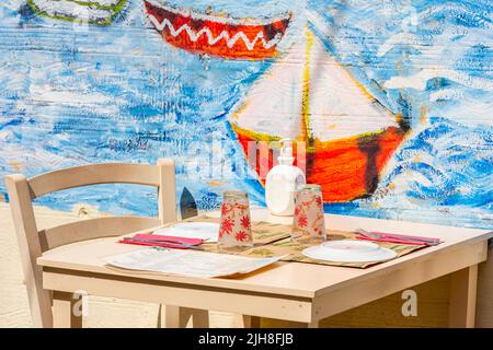 Tisch für zwei Personen in einer griechischen Taverne mit einem Segeljingboot-Wandgemälde an der Wand im Hintergrund, Tisch für zwei Personen, kretische Taverne, Segelyacht-Wandbild. Stockfoto