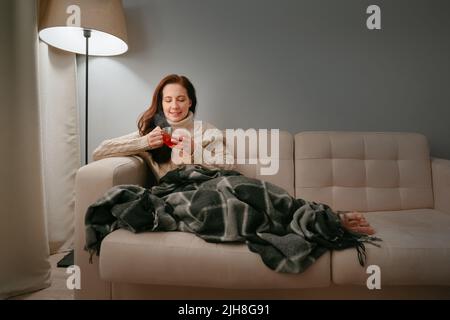 Die Frau, die sich in eine Decke eingehüllt hat, trinkt heißen Tee für Erkältungen. Stockfoto