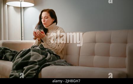 Eine Frau, die an einer Grippe erkrankt ist und eine Tasse heißen Hustentee auf ihn bläst. Stockfoto