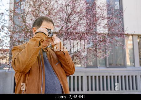 Eine Nahaufnahme eines älteren Mannes, der seine alte Filmkamera genießt und die Stadt vor dem Hintergrund eines blühenden rosa Kirschbaums fotografiert Stockfoto