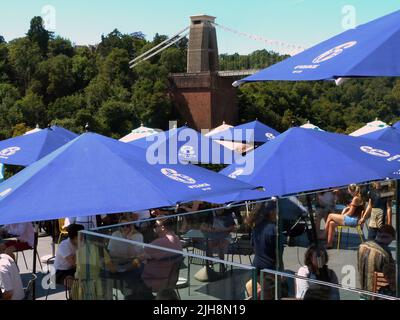 Die berühmte Clifton Suspension Bridge an einem sonnigen Sonntagnachmittag. Das ursprünglich von Isambard Kingdom Brunel entworfene und 1835 eröffnete Hotel erstreckt sich über die Avon-Schlucht über dem Fluss Avon in Clifton, Bristol. Es ist die bekannteste Struktur von Bristol und ist 365 Tage im Jahr geöffnet, mit einer Maut von £1,00, die mit einem Fahrzeug zu überqueren ist. Die Terrassenlage des Avon Gorge Hotels zieht einen konstanten Zustrom von Besuchern an, die den Blick auf dieses historische denkmalgeschützte Gebäude aus dem Jahr 1 genießen, während sie Erfrischungen unter schattigen Tischbrolles von diesem beliebten Aussichtspunkt genießen. Stockfoto