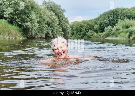 Ältere glückliche lachende Frau genießt es, in einem ruhigen Fluss auf dem Land zu schwimmen. Stockfoto