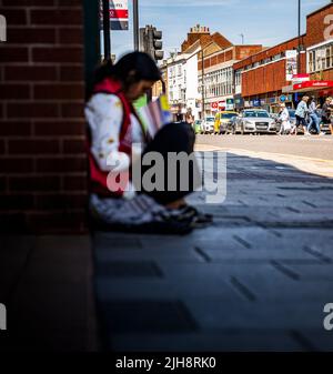 Grantham, Lincolnshire, Großbritannien – Eine junge asiatische Frau saß auf dem Straßenweg und verkaufte Kopien von The Big Issue, während die Leute an ihr vorbeigingen und sie ignorierten Stockfoto