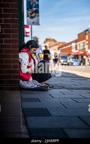 Grantham, Lincolnshire, Großbritannien – Eine junge asiatische Frau saß auf dem Straßenweg und verkaufte Kopien von The Big Issue, während die Leute an ihr vorbeigingen und sie ignorierten Stockfoto