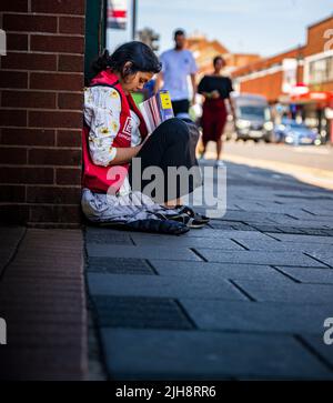 Grantham, Lincolnshire, Großbritannien – Eine junge asiatische Frau saß auf dem Straßenweg und verkaufte Kopien von The Big Issue, während die Leute an ihr vorbeigingen und sie ignorierten Stockfoto