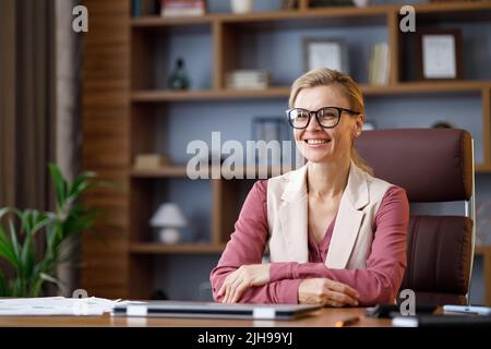 Porträt einer schönen blondie Frau, die die Kamera anschaut und lächelt. Erwachsene stilvolle selbstbewusste Geschäftsfrau trägt Brillen sitzen am Arbeitsplatz in Stockfoto