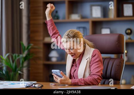 Glückliche Geschäftsfrau überrascht von den guten Nachrichten und macht ja Geste beim Blick auf Smartphone im Büro sitzen. Der Manager genießt den Erfolg Stockfoto