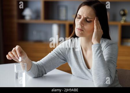 Verärgerte junge Frau, die unter Kopfschmerzen leidet, mit löslichem Medikament zur Schmerzlinderung. Erschöpfte Frau mit schmerzhaften Empfindungen. Migräne, hoher Blutdruck Stockfoto