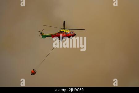 Mittelgroßer Hubschrauber von INFOCA, der einen Wassereimer trägt, um auf ein Waldfeuer zu fallen, Mijas, Spanien. Stockfoto