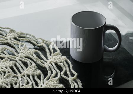 Porzellanbecher und weißes Mittelstück gehäkelt auf einem Glastisch. Handgestrickte Tischdecke für den Speisesaal mit Herzformen. Weißer Topf auf einem Glas ta Stockfoto