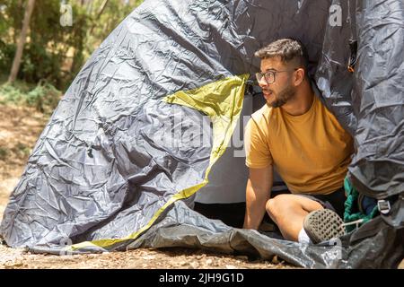 Junge in einem gelben T-Shirt und einer Brille, die in den Eingang seines dunkelgrünen Zeltes in der Mitte der Natur gucken. Konzept, um frische Luft zu genießen Stockfoto