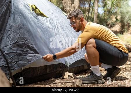 Junger Mann in gelbem T-Shirt mit Brille in einem Wald, der sein dunkelblaues Zelt für das Campen im Wald vorbereitet. Stockfoto