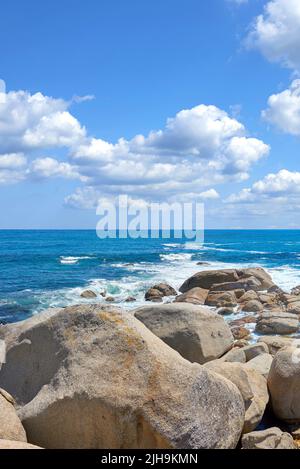 Copyspace auf See mit einem wolkigen blauen Himmel Hintergrund und felsigen Küste in Camps Bay, Kapstadt, Südafrika. Felsbrocken an einem Strand Ufer über eine majestätische Stockfoto