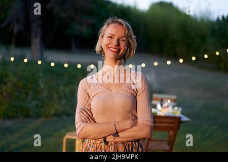 Porträt einer skandinavisch blonden Frau mittleren Alters, die die Kamera anschaut. Lächelnde nordische Frau im Hinterhof oder in einem Restaurant. Stockfoto