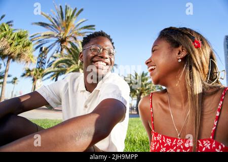 Fröhliches junges afrikanisches Paar, das bei ihrem ersten Date im Park in einer romantischen Outdoor-Liebhaberszene lacht und Spaß hat, auf dem Gras sitzt und redet Stockfoto