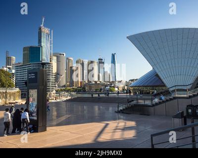 Touristen auf dem Vorplatz des Sydney Opera House mit dem Circular Quay und den CBD-Wolkenkratzern im Hintergrund. An einem sonnigen Wintertag. Stockfoto