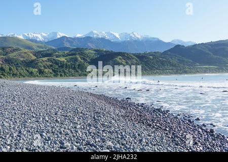 Stony Mangamaunu, Strand in der Nähe von Kaikoura mit Wellenreiten und Surfern vor dem Hintergrund der Küstenkette mit schneebedeckten Bergen. Stockfoto