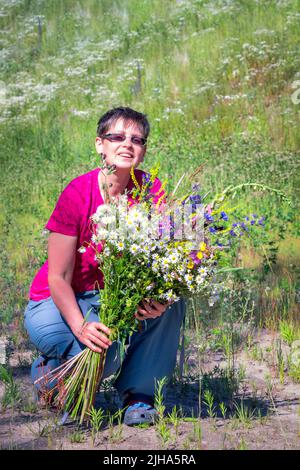 Lächelnde Frau mit einem riesigen Blumenstrauß, während sie im Gras sitzt. Stockfoto