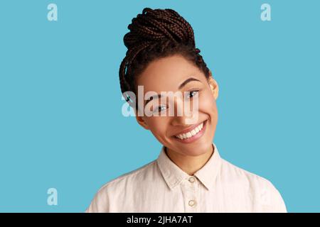 Portrait of Pleasant adorable Frau mit schwarzen Dreadlocks angenehme Optik, glücklicher Ausdruck, neigt den Kopf mit toothy Lächeln, trägt weißes Hemd. Innenaufnahme des Studios isoliert auf blauem Hintergrund. Stockfoto