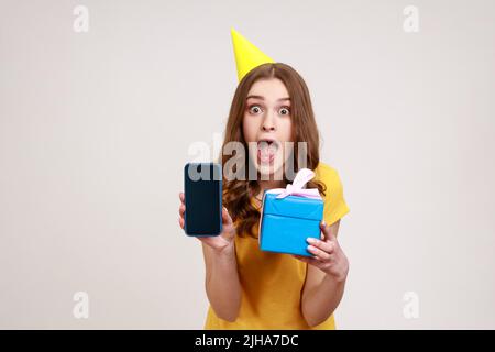 Unglaublicher Geburtstagsbonus für mobile Benutzer. Porträt von schockierten Teenager-Mädchen im Geburtstagskegel mit Geschenkbox und Handy mit leerem Display. Innenaufnahme des Studios isoliert auf grauem Hintergrund. Stockfoto