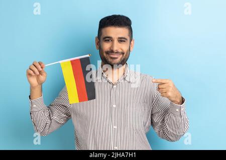 Porträt eines erfreuten Mannes mit Bart, der auf die deutsche Flagge zeigt, zur Feier des Deutschlandtages - 3.. Oktober, in weißem Hemd, in gestreiftem Hemd. Innenaufnahme des Studios isoliert auf blauem Hintergrund. Stockfoto