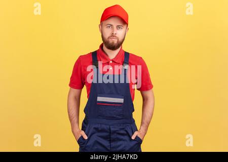 Porträt eines bärtigen Arbeiters in blauer Uniform und roter Mütze, der mit Händen in den Taschen steht und mit ernstem Gesichtsausdruck auf die Kamera blickt. Innenaufnahme des Studios isoliert auf gelbem Hintergrund. Stockfoto