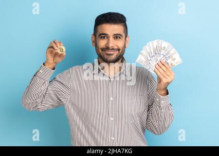 Ein glücklicher Geschäftsmann, der Dollarscheine und goldene Münzen der Kryptowährung zeigt, Geld in digitale Bitcoins überführt und ein gestreiftes Hemd trägt. Innenaufnahme des Studios isoliert auf blauem Hintergrund. Stockfoto