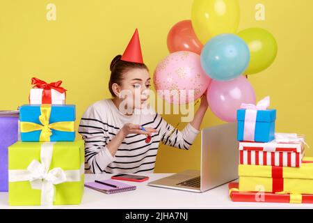 Dankbare Frau, die am Arbeitsplatz sitzt, umgeben von Geschenkboxen und Ballons, Luftküsse auf den Laptop-Bildschirm schickt und den Geburtstag per Videoanruf feiert. Studio-Innenaufnahme isoliert auf gelbem Hintergrund Stockfoto