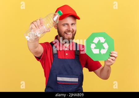 Porträt eines bärtig lächelnden Handwerkers, der stand und Recycling-Schild und Plastikflasche hielt, grün dachte, Overalls und rote Mütze trug. Innenaufnahme des Studios isoliert auf gelbem Hintergrund. Stockfoto
