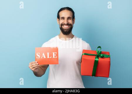 Boni beim Shopping am Black Friday. Porträt eines Mannes in weißem T-Shirt mit Aufschrift und Geschenkbox, der mit einem Lächeln auf die Kamera blickt. Innenaufnahme des Studios isoliert auf blauem Hintergrund. Stockfoto