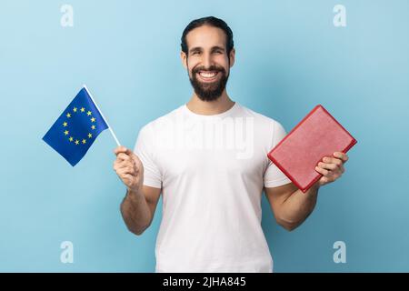 Porträt eines zufriedenen, lächelnden bärtigen Mannes mit weißem T-Shirt, der ein Buch und eine Europaflagge hält, Bildung im Ausland, und mit einem dutzenden Lächeln auf die Kamera blickt. Innenaufnahme des Studios isoliert auf blauem Hintergrund. Stockfoto