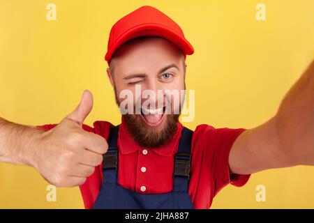 Zufriedener lächelnder Arbeiter in blauer Uniform, der Selfie macht, die Kamera winkend anschaut und den Daumen nach oben zeigt, aus der Sicht des Fotos. Innenaufnahme des Studios isoliert auf gelbem Hintergrund. Stockfoto