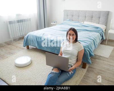 Weißer Roboter-Staubsauger, der den Boden reinigt, während die Frau einen Laptop benutzt Stockfoto