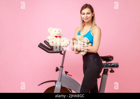 Porträt einer attraktiven, schönen, sportlichen Frau, die Teddybär hält, in der Nähe von Sportgeräten posiert, Heimtrainer, Sportstrumpfhose und Oberteil trägt. Innenaufnahme des Studios isoliert auf rosa Hintergrund. Stockfoto