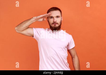 Ja, Sir. Porträt eines ernsthaften, strengen bärtigen Mannes, der als Soldat steht und einen Gruß gibt, bereit zur Bestellung, in einem rosa T-Shirt. Innenaufnahme des Studios isoliert auf orangefarbenem Hintergrund. Stockfoto