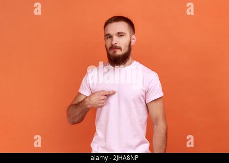 Porträt eines bärtigen Mannes, der sich zeigt und mit arrogantem egoistischem Ausdruck aussieht, sich erfolgreich und selbstwichtig fühlt und ein rosa T-Shirt trägt. Innenaufnahme des Studios isoliert auf orangefarbenem Hintergrund. Stockfoto