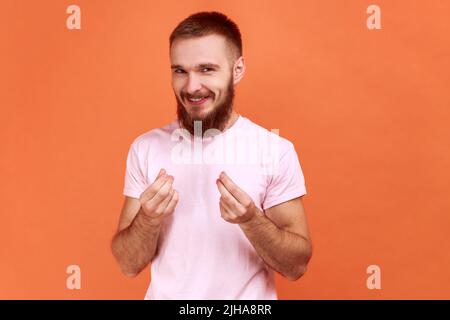 Porträt eines hübschen jungen bärtigen Mannes, der mit Geld oder italienischer Geste mit der Hand die Kamera anschaut und ein pinkes T-Shirt trägt. Innenaufnahme des Studios isoliert auf orangefarbenem Hintergrund. Stockfoto
