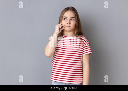 Portrait of adorable Little girl tragend gestreiftes T-Shirt mit Mystery Look machen Reißverschluss Geste zu schließen Mund, geheim zu halten, Zippen Lippen. Innenaufnahme des Studios isoliert auf grauem Hintergrund. Stockfoto