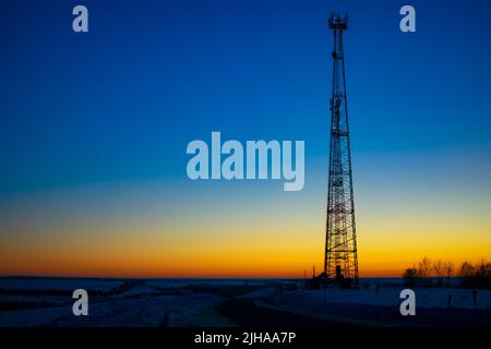Nachtansicht eines Metallfernsehturms auf einem Berg über einem Dorf in der Nähe einer verschneiten Autostraße im Winter vor einem dunklen, bläulich klaren Himmel Stockfoto