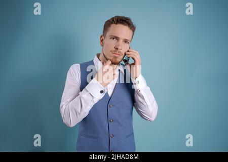 Serious jungen bärtigen Geschäftsmann in klassischen Anzug, der auf blauem Hintergrund Studios Porträt positioniert ist. Karriereerfolg Reichtum Geschäftskonzept. Auf den Kopierort ausfließen. Mobiltelefon wird gehalten Stockfoto
