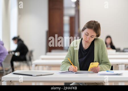 Konzentrierte reife Studentin, die Smartphone für die Prüfungsvorbereitung verwendet und den zweiten Abschluss macht Stockfoto