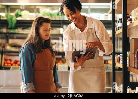 Glückliche Lebensmittelmanagerin, die ein digitales Tablet verwendet, während sie mit ihrem Mitarbeiter ein Gespräch führt. Weibliche Ladenbesitzerin, die eine Frau mit Down-Syndrom tr Stockfoto