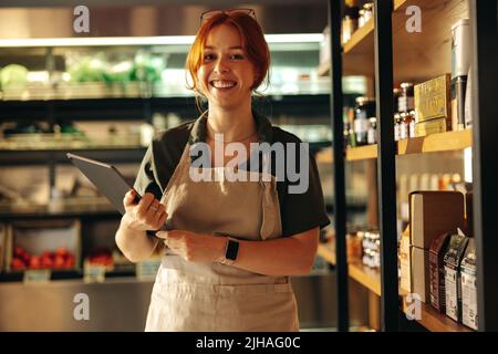 Glücklicher junger Besitzer eines Lebensmittelladens, der die Kamera anlächelt, während er ein digitales Tablet hält. Fröhliche weibliche Unternehmerin, die ein erfolgreiches kleines Unternehmen betreibt Stockfoto