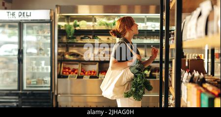 Weibliche Shopper, die Lebensmittel aus einem Regal auswählen, während sie eine Tasche mit Gemüse in einem Lebensmittelgeschäft tragen. Junge Frau macht ein paar Lebensmitteleinkäufe in Stockfoto