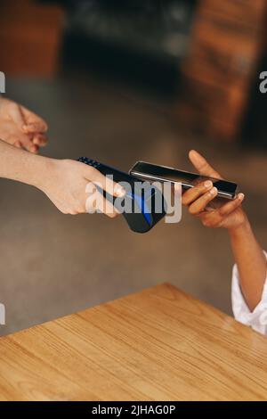 Cafe-Kundin scannt ihr Smartphone auf einem Kreditkartenrechner, um ihre Rechnung zu bezahlen. Nicht erkennbare Frau, die eine bargeldlose und kontaktlose Transaktion usin Stockfoto