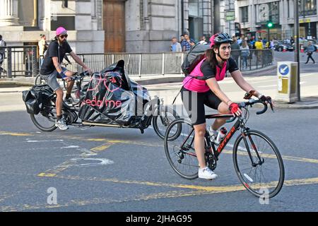 Radfahrerin mit Rucksack-Fahrrad mit Fallgriff über die Kreuzungsstraße der City of London, gefolgt von einem Mann auf dem langen Lastenrad England Großbritannien Stockfoto