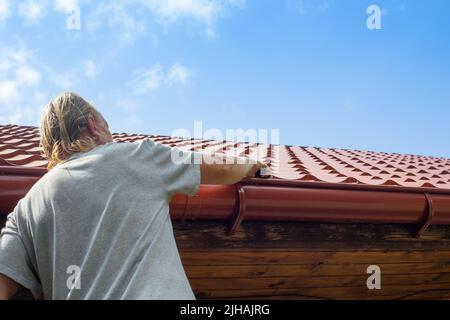 Ein Mann reinigt die Rinne eines Abflussrohres von trockenen Blättern und Trümmern. Vermeidung von Stauungen und Bruch des Abflusses der Rinne. Stockfoto